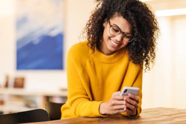 Gözlüklü mutlu bir Afro-Amerikan kadının masada otururken akıllı telefon kullanması.