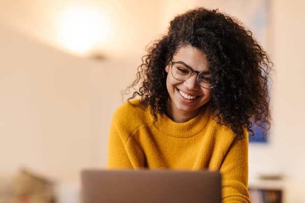 Image of cheerful african american woman in eyeglasses using laptop while sitting at living room