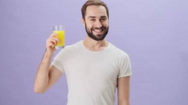 Beyaz t-shirt giyen genç bir adam bir bardak portakal suyu gösteriyor ve gri arka planda izole bir şekilde baş parmak hareketi yapıyor.
