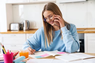 Gözlüklü neşeli bir kadının evde egzersiz kitaplarıyla çalışırken cep telefonuyla konuşması.
