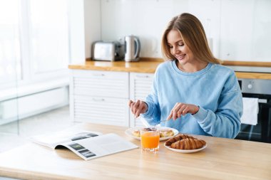 Modern mutfakta kahvaltı ederken gülümseyen, sarı saçlı neşeli genç bir kadın resmi.