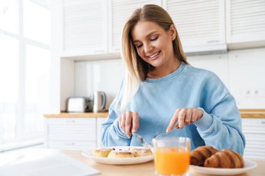 Modern mutfakta kahvaltı ederken gülümseyen, sarı saçlı neşeli genç bir kadın resmi.
