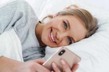 Yatakta yattıktan sonra gülümseyen ve cep telefonu kullanan güzel bir kadının yakın plan fotoğrafı.