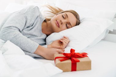 Hediye kutusuyla uzanmış, yatakta uyuyan, sarı saçlı, hoş, beyaz bir kadın resmi.