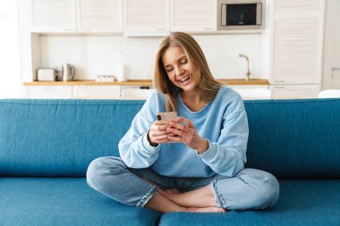 Akıllı telefon kullanan ve evdeki kanepede oturup gülen neşeli genç bir kadın görüntüsü.