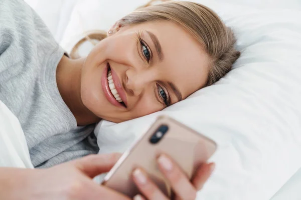 Image closeup of cute joyful woman smiling and using cellphone while lying after sleep in bed