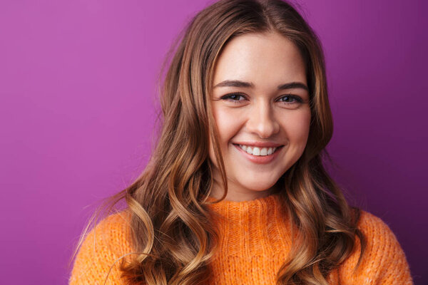 Портрет милой веселой девушки в свитере, стоящей изолированно на фиолетовом фоне, смотрящей в камеру
