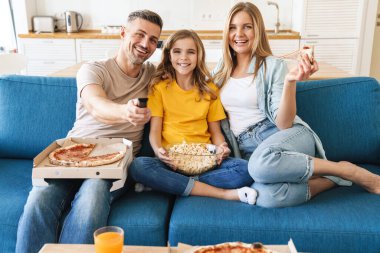 Evde televizyon izlerken patlamış mısır ve pizza yiyen neşeli beyaz bir ailenin fotoğrafı.