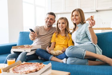 Evde televizyon izlerken patlamış mısır ve pizza yiyen neşeli beyaz bir ailenin fotoğrafı.