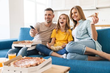 Evde televizyon izlerken patlamış mısır ve pizza yiyen neşeli beyaz bir ailenin fotoğrafı.