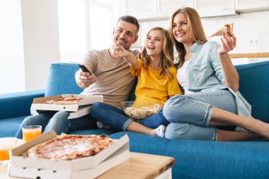 Evde televizyon izlerken patlamış mısır ve pizza yiyen neşeli beyaz bir ailenin fotoğrafı.