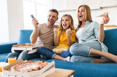 Heyecanlı beyaz bir ailenin evde televizyon izlerken patlamış mısır ve pizza yediği fotoğraf.