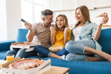 Heyecanlı beyaz bir ailenin evde televizyon izlerken patlamış mısır ve pizza yediği fotoğraf.