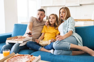 Evde televizyon izlerken patlamış mısır ve pizza yiyen korkmuş beyaz bir ailenin fotoğrafı.