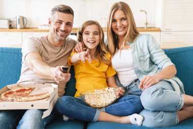 Evde televizyon izlerken patlamış mısır ve pizza yiyen neşeli beyaz bir ailenin fotoğrafı.