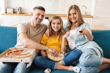 Evde televizyon izlerken patlamış mısır ve pizza yiyen neşeli beyaz bir ailenin fotoğrafı.