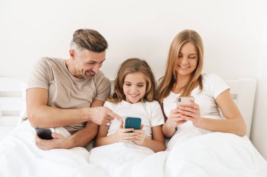 Uykudan sonra beyazlar içinde kötü bir şekilde otururken gülümseyen ve cep telefonu kullanan neşeli beyaz aile fotoğrafı.