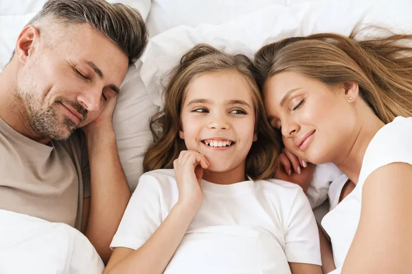 Photo of joyful caucasian girl laughing while lying with sleeping parents in white bad