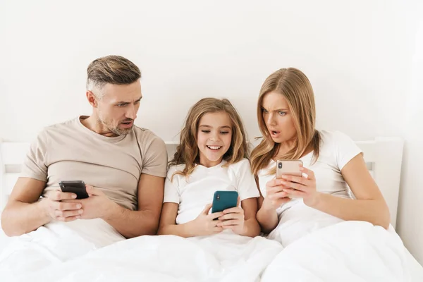 Photo of young caucasian family expressing shock and using cellphones while sitting in white bad after sleep