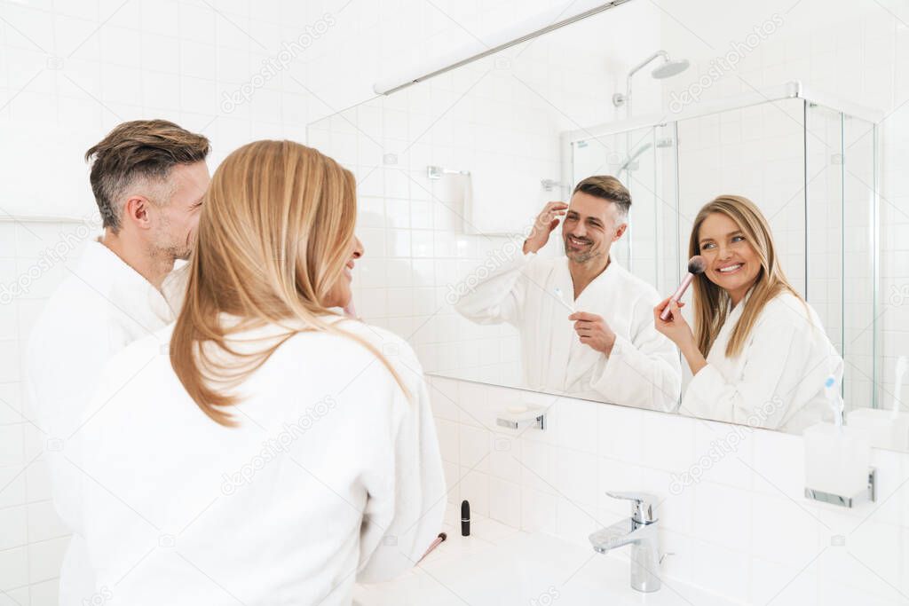Photo of happy caucasian couple wearing white bathrobes smiling and looking at mirror in bathroom