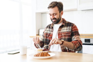 Mutfakta kahvaltı ederken gülümseyen ve pasta yiyen neşeli sakallı adamın portresi.