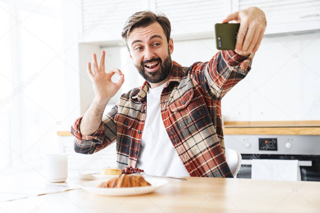 Portrait of smiling bearded man taking selfie on cellphone and showing ok sign while having breakfast at home