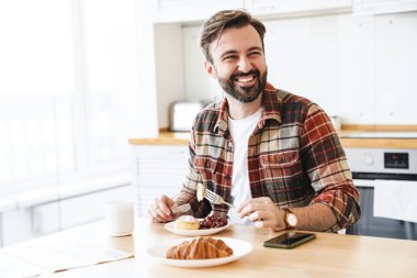 Mutfakta kahvaltı ederken gülümseyen ve pasta yiyen neşeli sakallı adamın portresi.