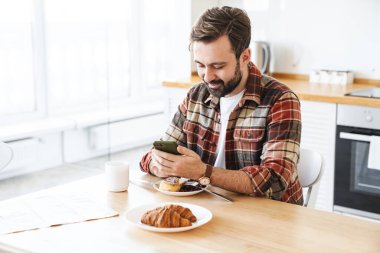 Evde kahvaltı yaparken cep telefonu kullanan ve gülümseyen neşeli sakallı adamın portresi.