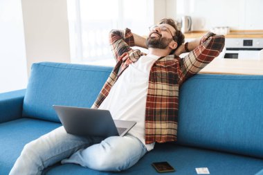 Evdeki kanepede otururken gülümseyen ve dizüstü bilgisayar kullanan sakallı genç adamın portresi.