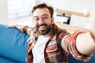 Evdeki kanepede otururken gülümseyen ve selfie çeken genç bir adam resmi.