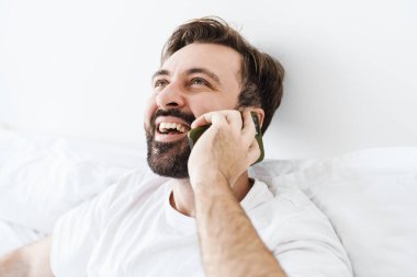 Evde yatarken gülümseyen ve cep telefonuyla konuşan genç, tıraşsız beyaz adam.