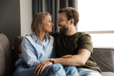 Evdeki kanepede otururken gülümseyen ve sarılan neşeli, romantik beyaz bir çiftin fotoğrafı.