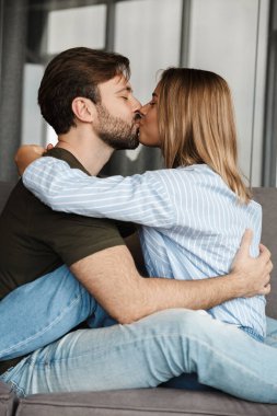 Mutlu, romantik, beyaz bir çiftin evdeki kanepede oturup öpüşürken ve sarılırken çekilmiş bir fotoğrafı.