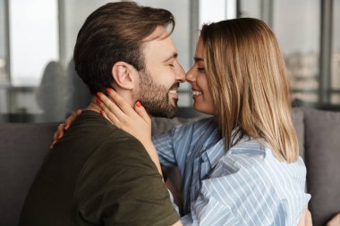 Evdeki kanepede otururken gülümseyen ve sarılan neşeli, romantik beyaz bir çiftin fotoğrafı.