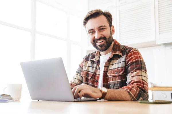 一个满脸胡须的快乐男人坐在家里的桌子边 一边用笔记本电脑一边笑的画像 — 图库照片
