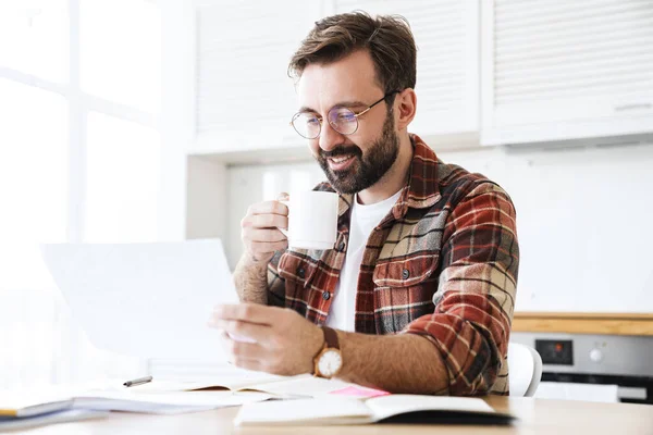 年轻留着胡子的男人戴着眼镜边喝茶边在家里做笔记的画像 — 图库照片