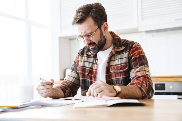 Portrait of young bearded displeased man wearing eyeglasses working with notes at home