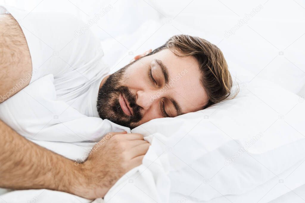 Image of young unshaven caucasian man sleeping alone in bed with white linen at home
