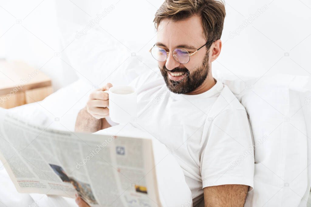 Image of young unshaven caucasian man drinking coffee and reading newspaper while lying in bed at home