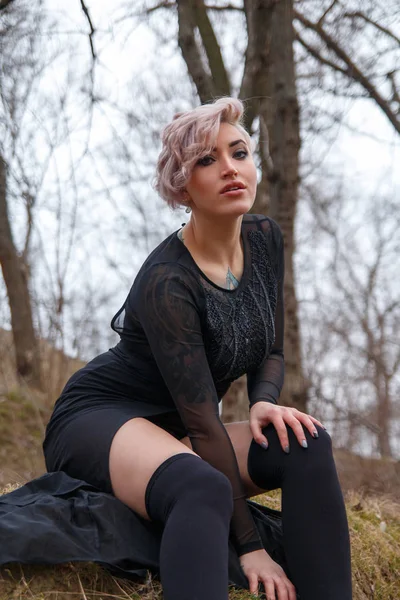 Retrato de uma jovem sentada na floresta em vestido preto e — Fotografia de Stock