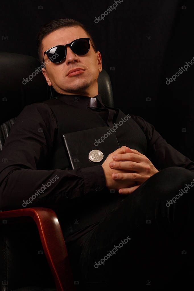 Modern priest holding and crypto bible with bitcoin logo sitting in the armchair