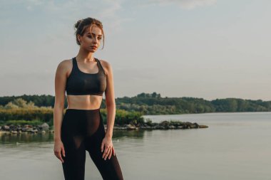 Genç seksi atletik kız gün batımında nehrin kenarında duruyor.