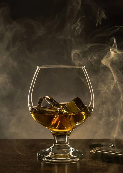 Glass with Alcohol and Ice Cubes on Wooden table. Smoke and Copy Space in Background.