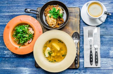 Öğle Yemeği: tavada et ve otlar, sarı çorba, salata sebze ve otlar, kahve ve çatal bıçak takımı, mavi kot arka plan üzerinde. Yukarıdan görüntülemek