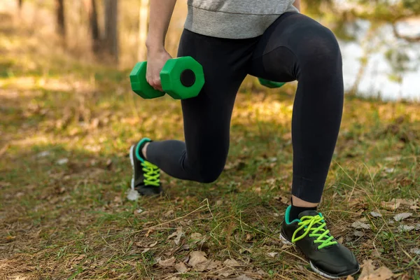 Girl in sports pants and hoodie, crouching with green dumbbells