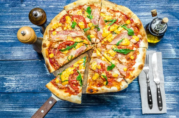 Round pizza with meat, tomatoes, corn and herbs, cutlery and spices, on a blue denim background