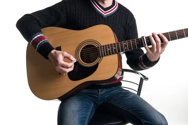 Gitarrist i jeans och en svart tröja spelar en akustisk gitarr med ett reglage som sitter på en stol i mitten av ramen på en vit bakgrund. Horisontell ram. — Stockfoto