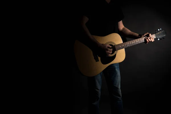 Il chitarrista in jeans suona una chitarra acustica, sul lato destro del telaio, su uno sfondo nero. Telaio orizzontale — Foto Stock