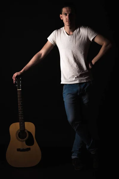 Κιθαρίστας, μουσική. Ένας νεαρός άνδρας στέκεται με μια ακουστική κιθάρα πάνω σε μαύρο φόντο απομονωμένες. Κατακόρυφο πλαίσιο — Φωτογραφία Αρχείου