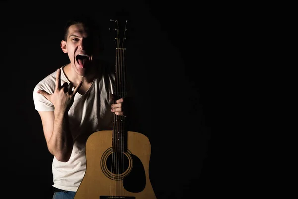 Гітарист, музика. Молодий чоловік стоїть з акустичною гітарою і показує язик і пальці на чорному ізольованому тлі. Горизонтальна рамка — стокове фото